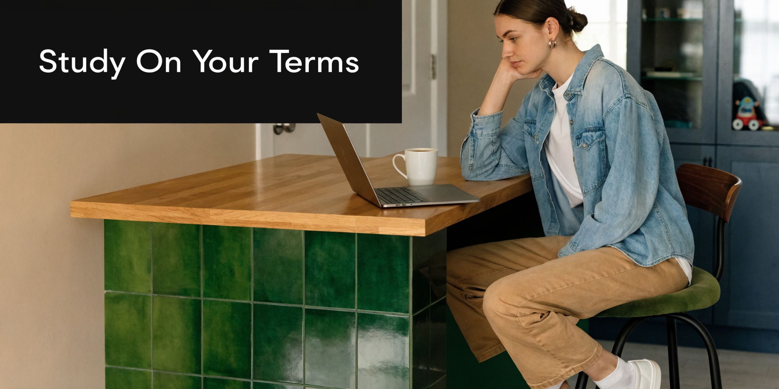 A young woman studying on her laptop at a kitchen counter with a cup of coffee nearby.
