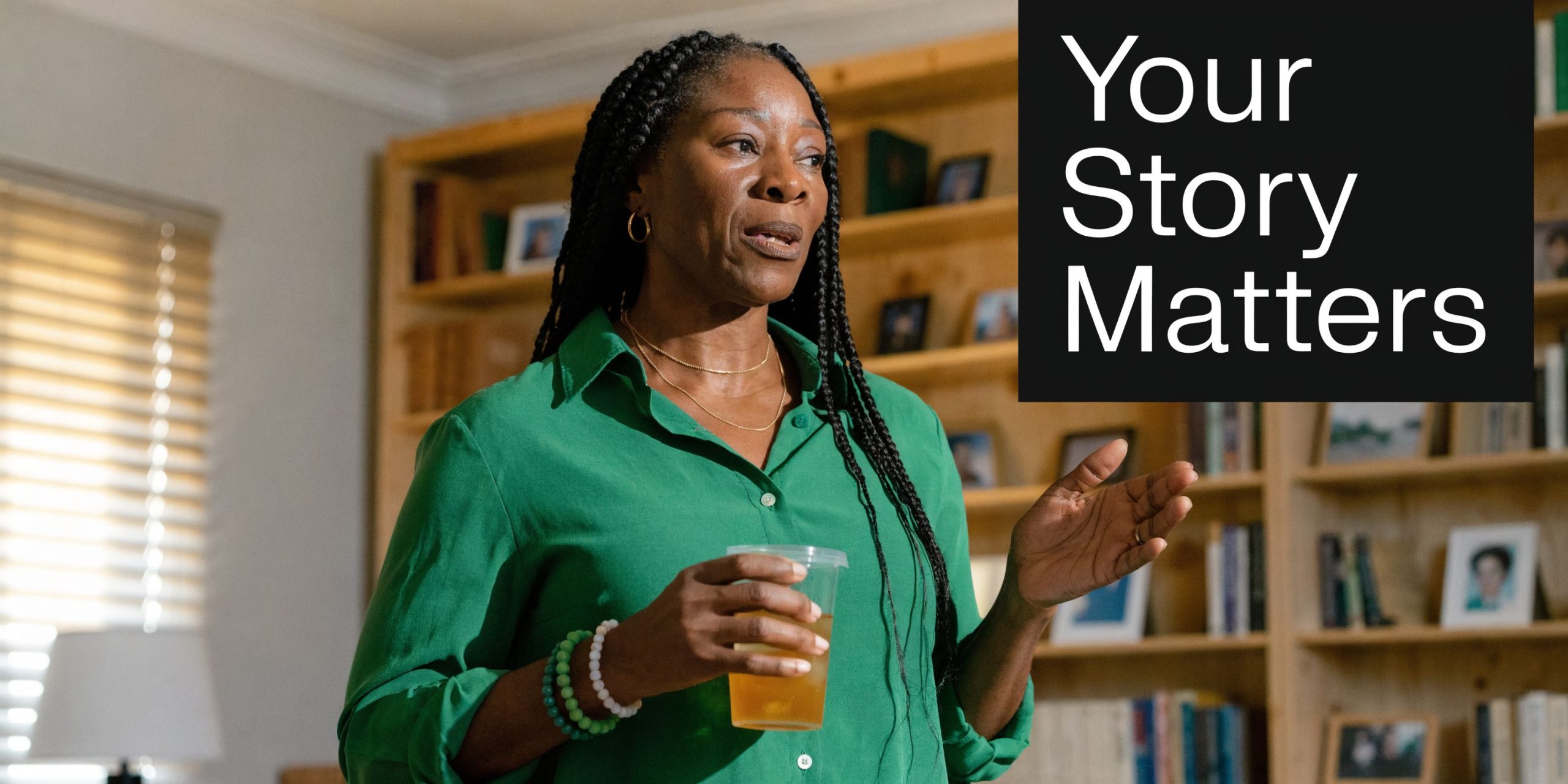 A middle aged Black woman wearing a green shirt holding a drink while speaking in a room.