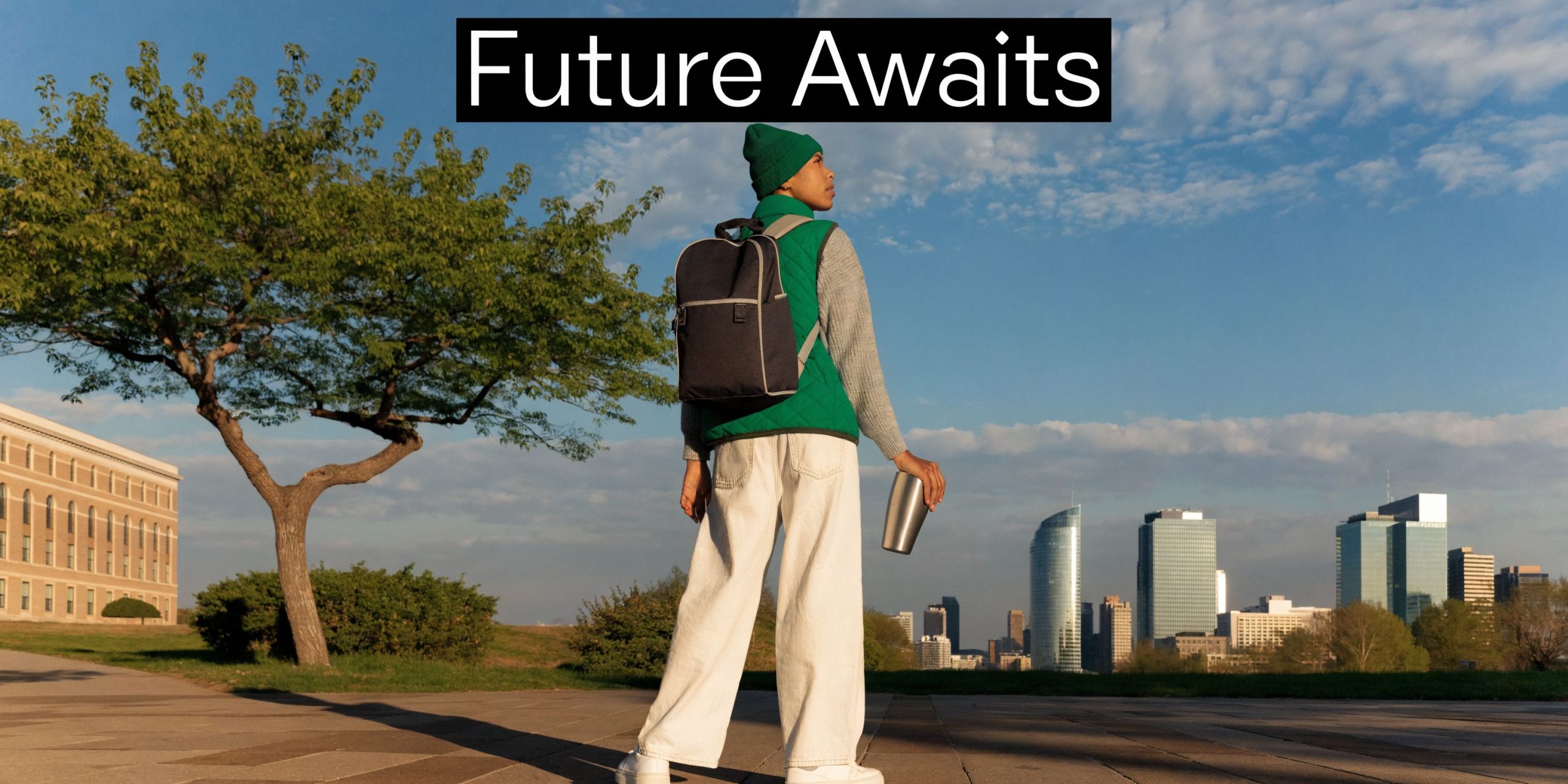 A person standing on a path facing a city skyline during sunset, wearing a backpack and hat.