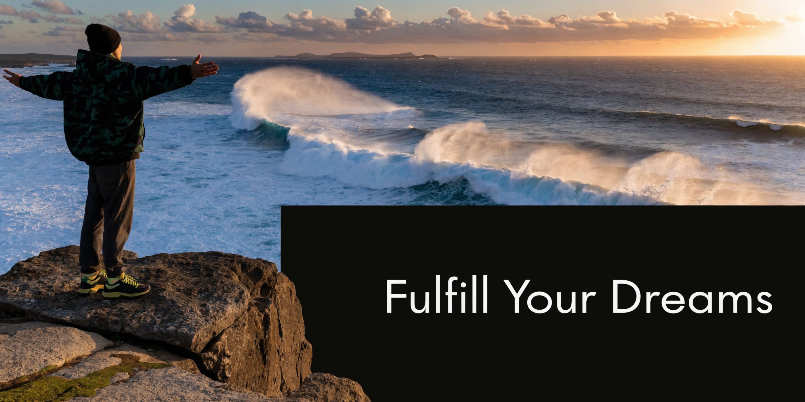 A person standing on a rocky cliff overlooking the ocean with arms wide open at sunset.