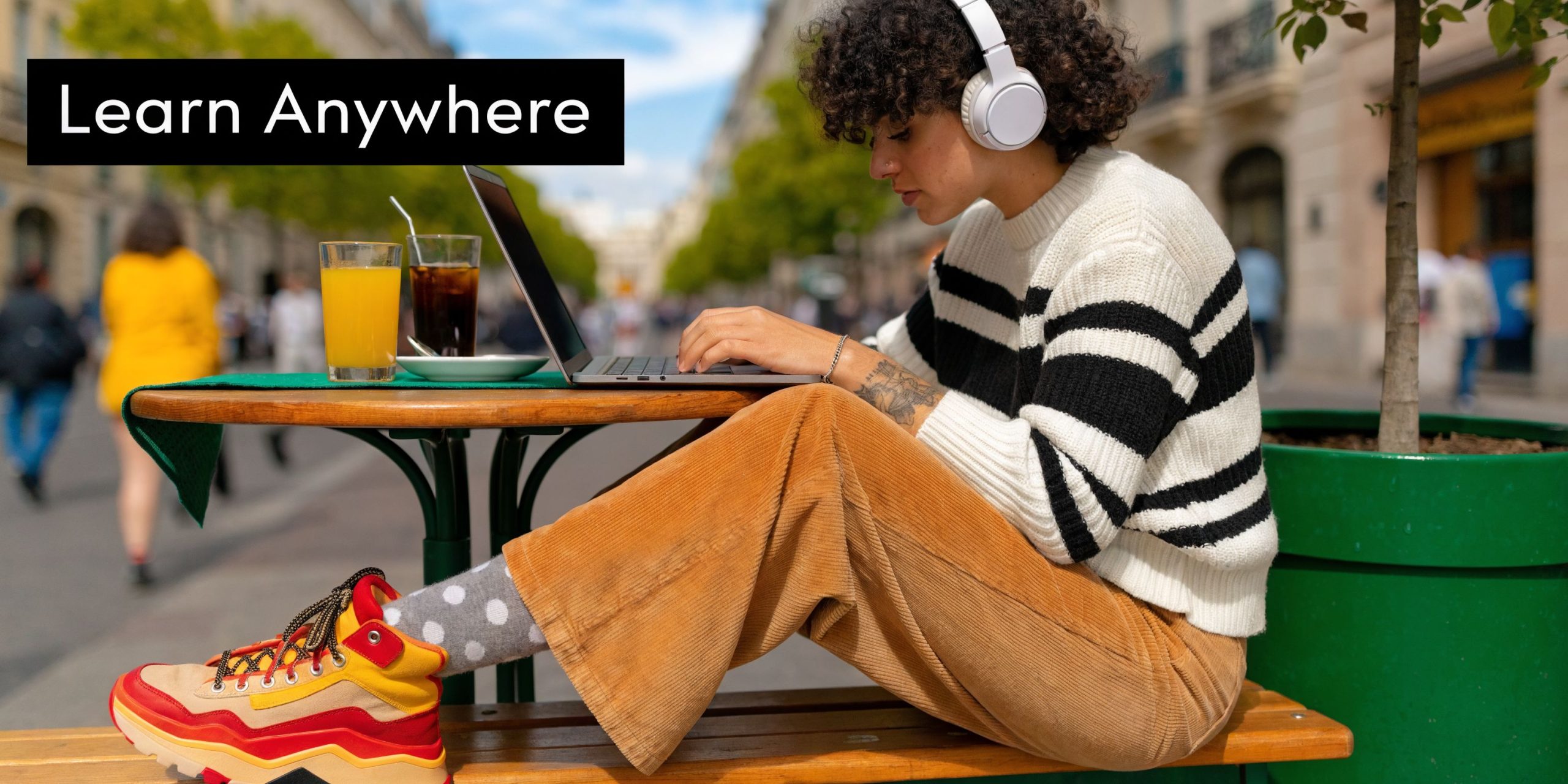 A person with curly hair wearing headphones using a laptop at an outdoor table in a city.