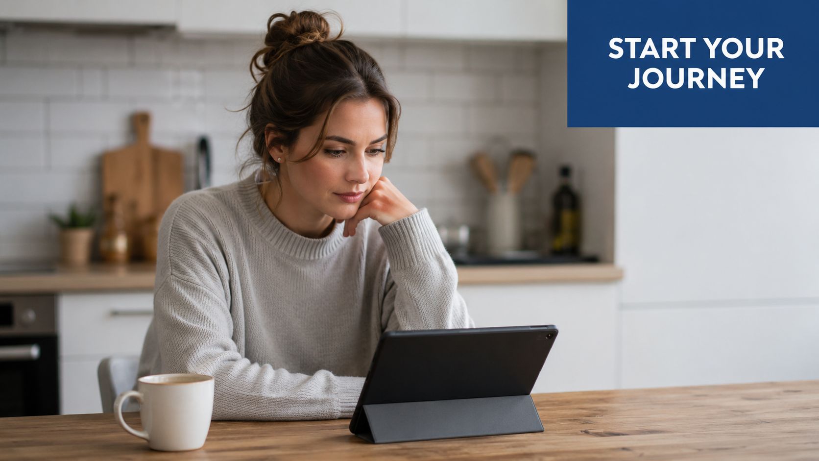 A woman sits at a wooden table in a kitchen, looking thoughtfully at her digital tablet device.